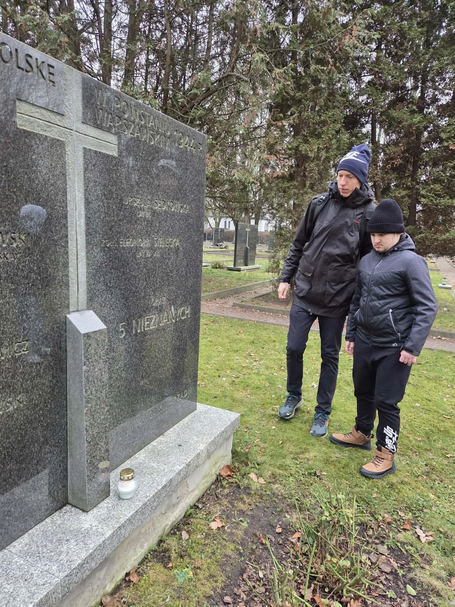 Uczeń wraz z nauczycielem przy mogile na Cmentarzu Powstańców Warszawy przy zapalonym zniczu. 