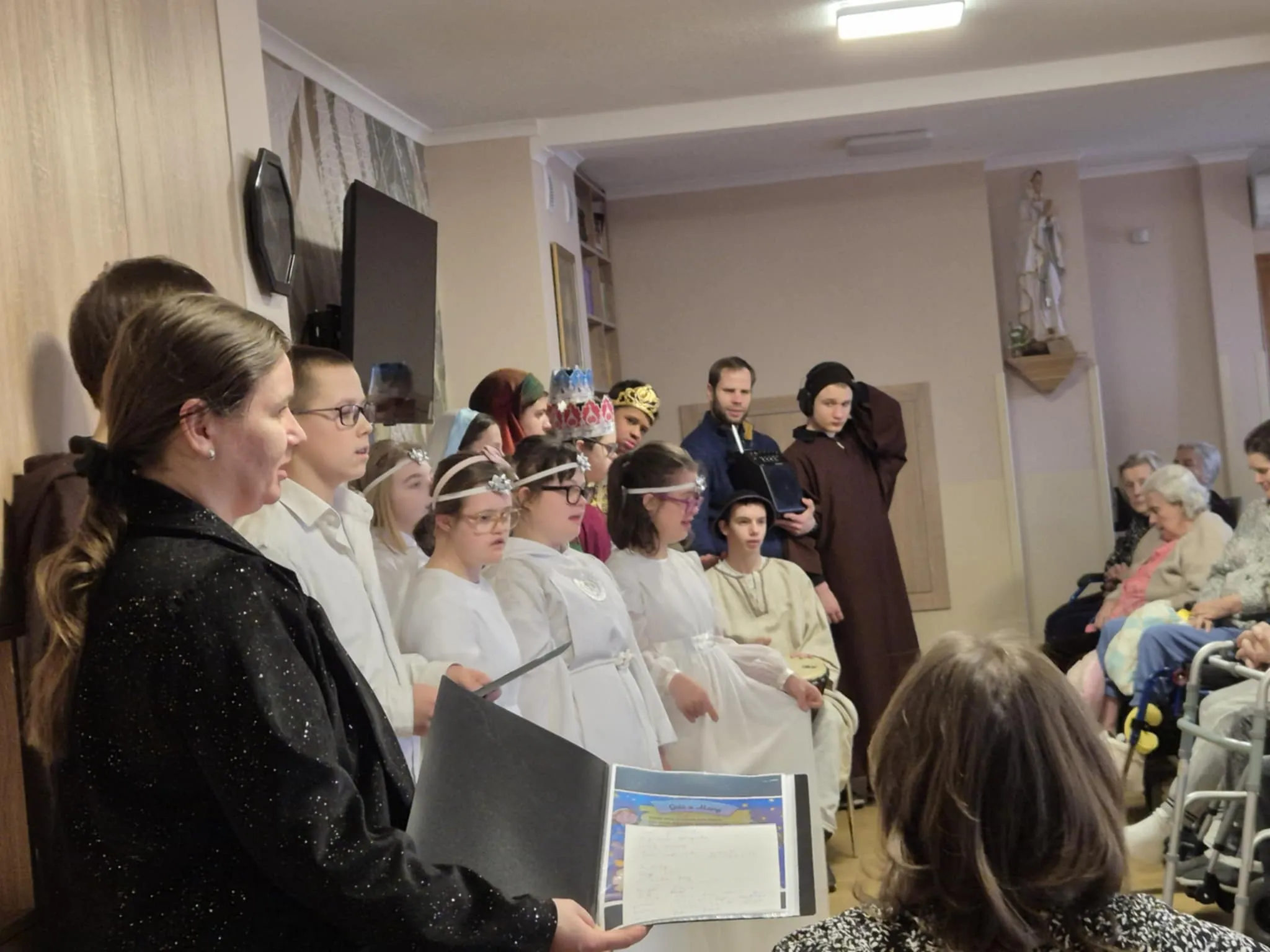 Fragment występu uczniów w zakładzie opiekuńczym. Uczniowie przebrani są za postacie  związane z narodzeniem  Jezusa.  Z prawej strony zdjęcia siedzi pani katechetka z gitarą oraz stoi  jedna z pan wychowawczyń szkoły.