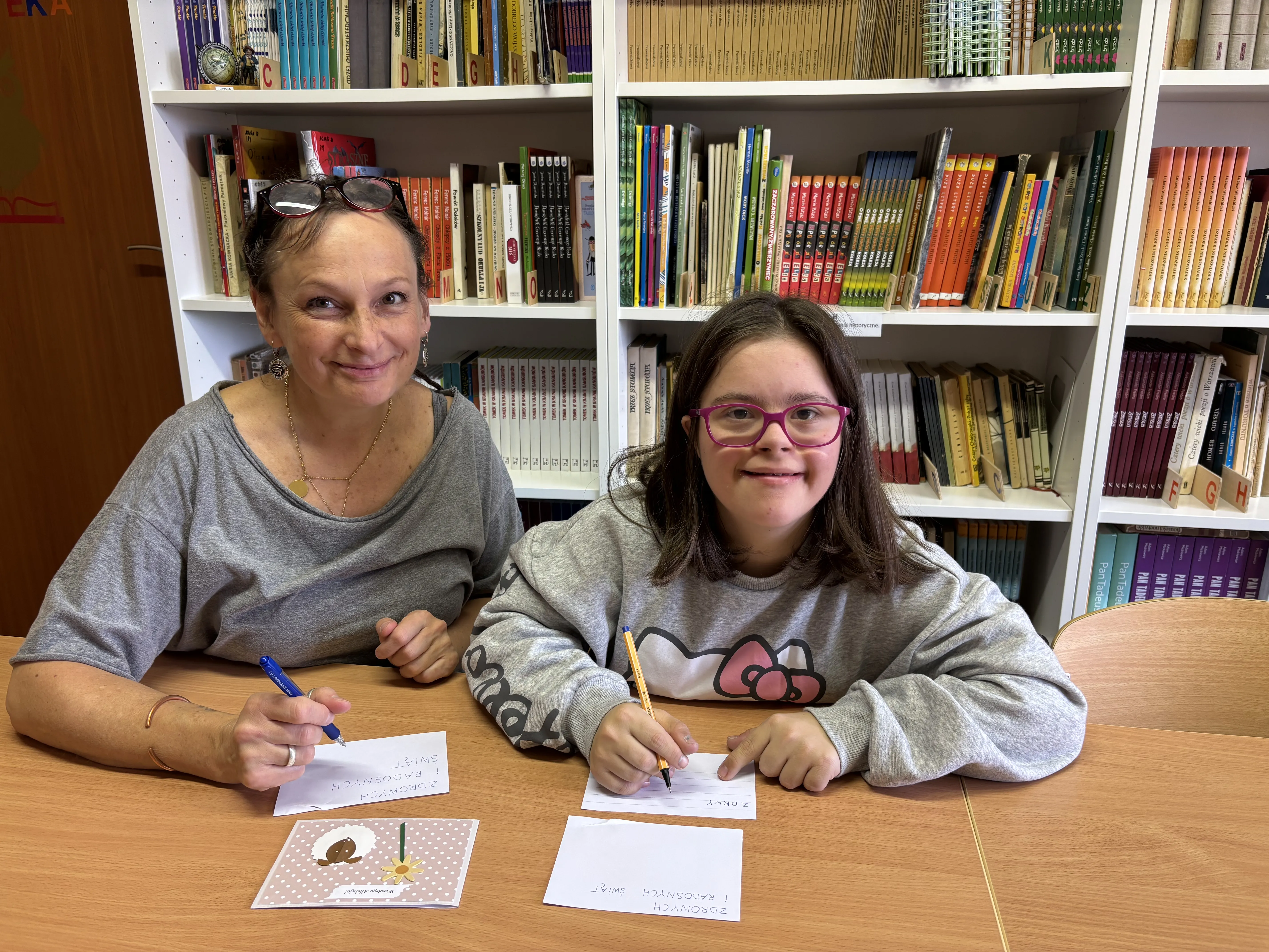 Uczennica klasy 7c prezentuje swoją kartkę świąteczną w bibliotece szkolnej. Towarzyszy jej nauczycielka logopedii.