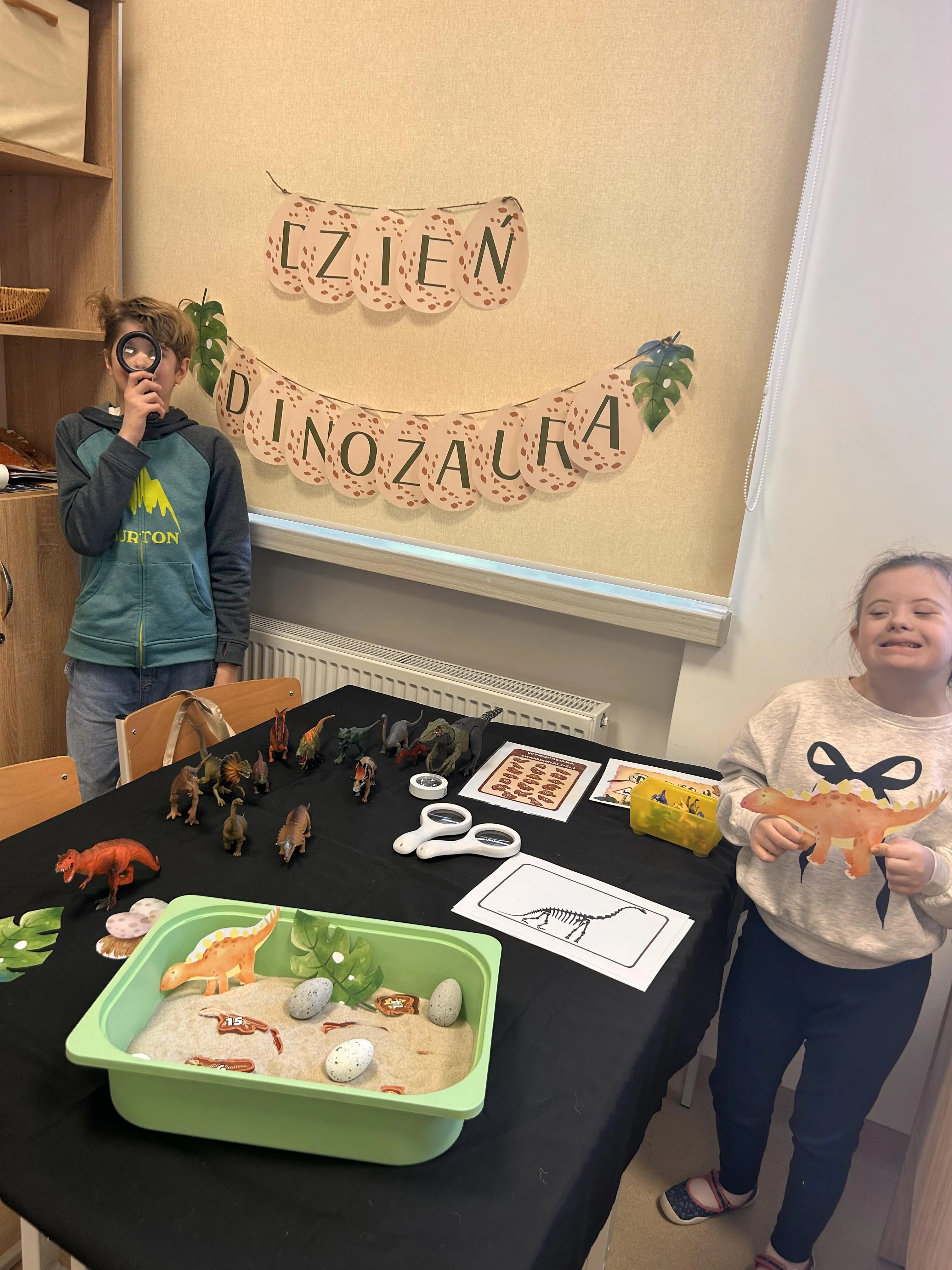 Uczeń i uczennica przy stole, na którym porozkładane są modele dinozaurów oraz plansze i przedmioty i materiały do eksperymentów.  Uczennica trzyma model dinozaura, a uczeń lupę.