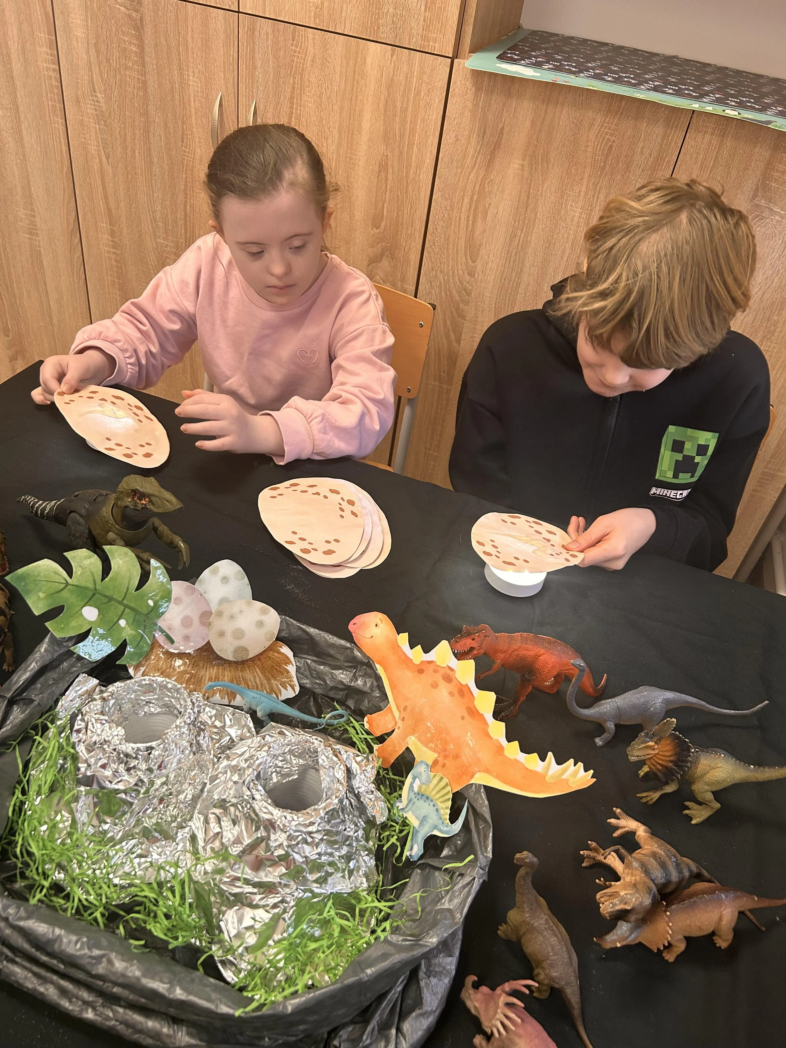 Uczeń i uczennica siedzą przy stoliku, na którym porozkładane są akcesoria związane z dinozaurami , przyglądają się planszom z różnymi śladami 