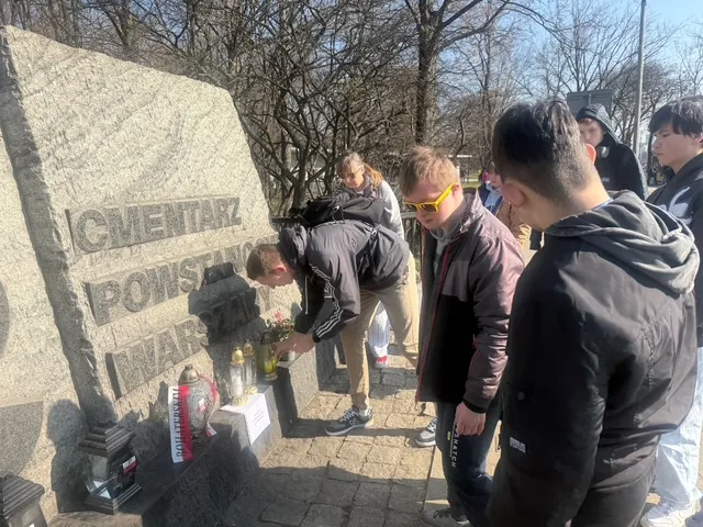 Grupa uczniów z obojgiem nauczycieli przy płycie cmentarnej z napisem Cmentarz Powstańców  Warszawy. Nauczyciele  na dole płyty ustawiają znicze . 