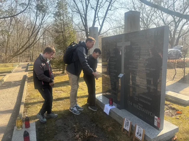 Dwoje uczniów wraz z nauczycielem przy mogile,  na której ustawione są dwa znicze