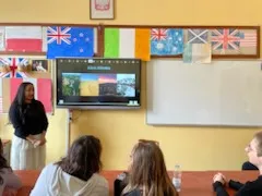 Na ścianie w klasie wisi włączona tablica multimedialna, obok stoi studentka .   W dolnej części zdjęcia widoczni są  uczniowie obserwujący tablicę. Na ścianie w klasie widoczne są rozwieszone  kartony z flagami różnych państw.