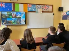 Na ścianie w klasie wisi tablica multimedialna.  W dolnej części zdjęcia widoczni są  uczniowie obserwujący tablicę. Na ścianie w klasie widoczne są rozwieszone  kartony z flagami różnych państw.