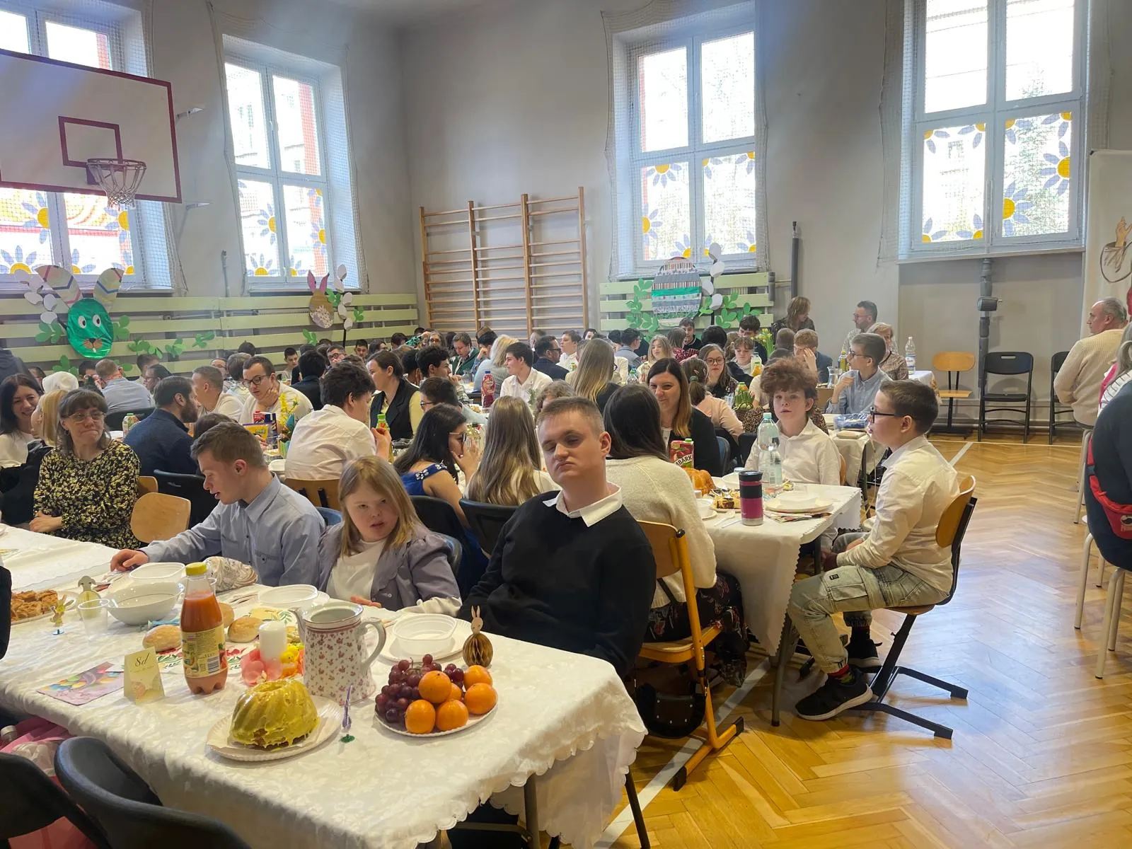 uczniowie i nauczyciele siedzacy przy stołach w sali gimnastyczne przy śniadaniu wielkancnym