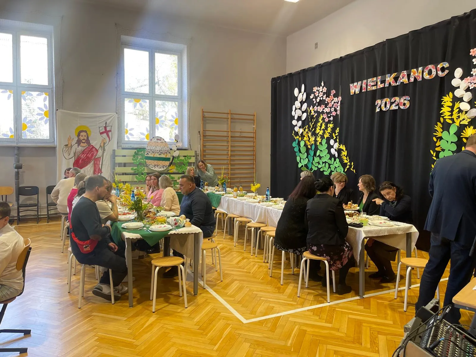 uczniowie i nauczyciele siedzacy przy stołach w sali gimnastyczne przy śniadaniu wielkancnym