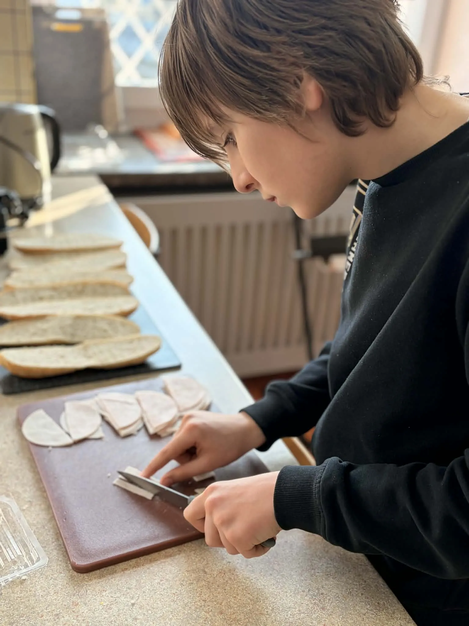 Na zdjęciu chłopiec stojący przy blacie kuchennym kroi na desce  wędlinę. Obok leżą  pokrojone kawałki wędliny oraz przekrojone bagietki.
