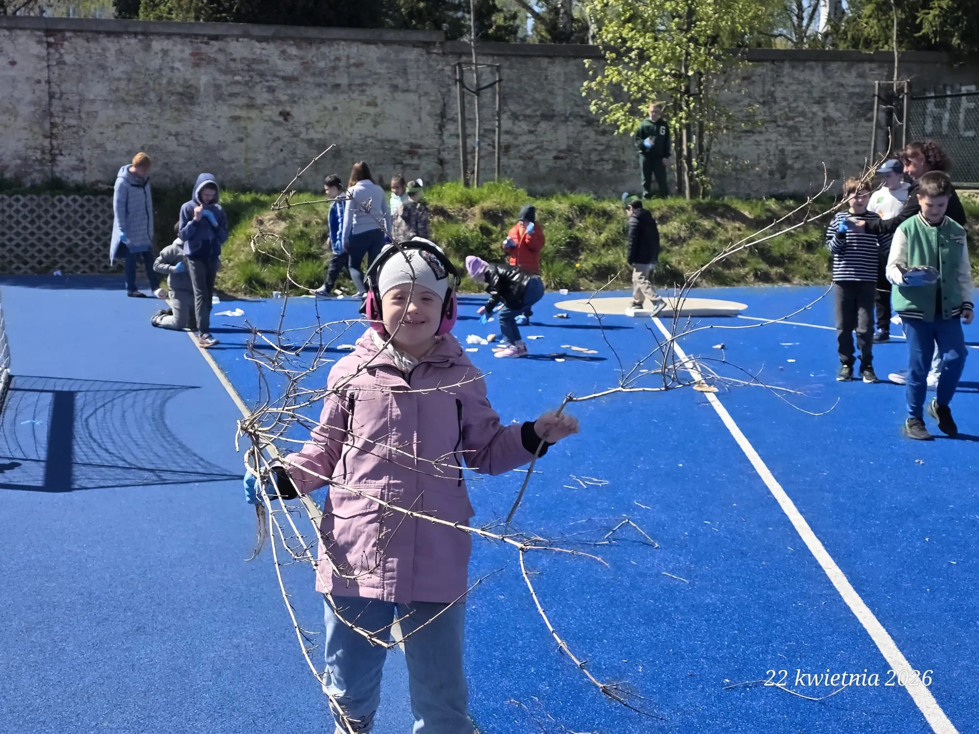Na pierwszym planie zdjęcia - widoczna uczennica, która trzyma w ręku suche gałązki. W głębi zdjęcia boisko szkolne  i zbierający śmieci  uczniowie.
