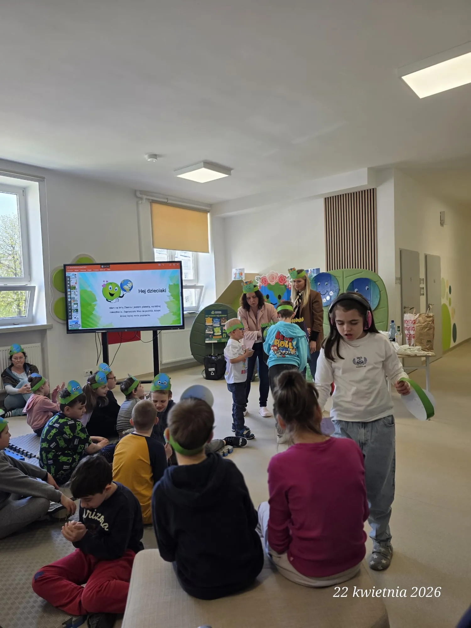 Na zdjęciu uczniowie w czapeczkach z wizerunkiem Ziemi lub zielonych opaskach. Część z nich siedzi na korytarzu, niektórzy stoją.  W głębi zdjęcia widoczna jest tablica multimedialna, w pobliżu niej  stoją 2 nauczycielki  oraz 2 uczniów. 